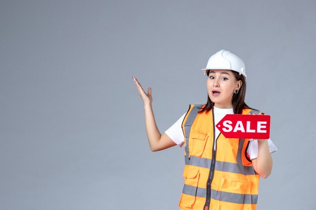 Front view of female builder holding red sale board on gray wall