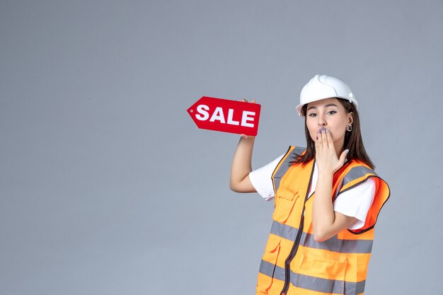 Front view of female builder holding red sale board on gray wall