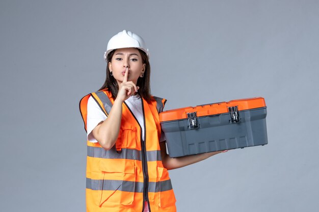 Front view of female builder holding tool case and asking to be silent on gray wall