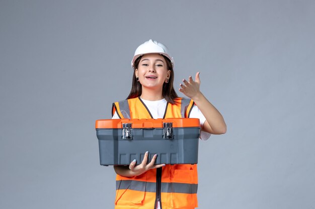 Front view of female builder holding tool case on gray wall