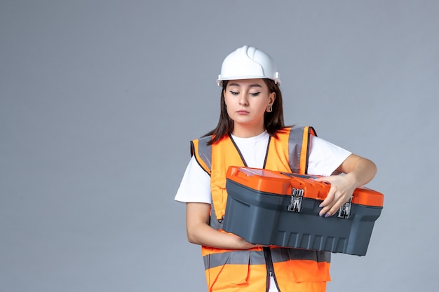 Front view of female builder holding tool case on gray wall