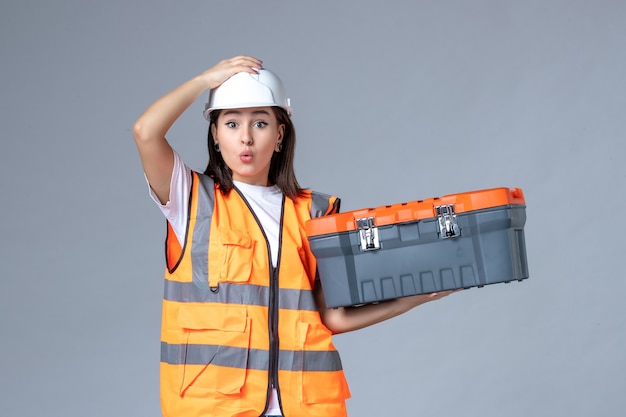 Front view of female builder holding tool case on gray wall