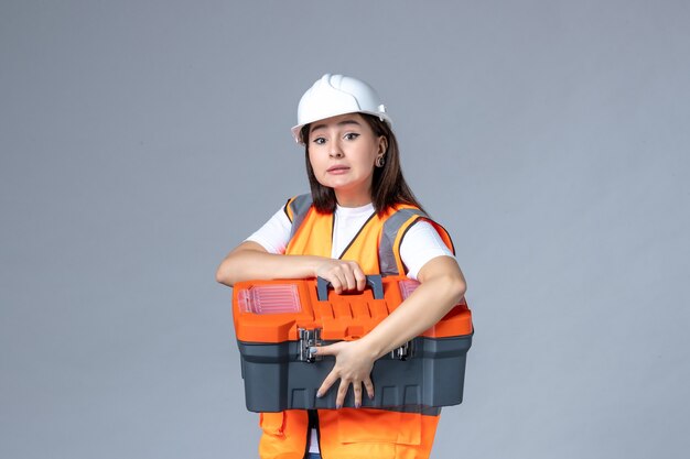 Front view of female builder holding tool case on gray wall