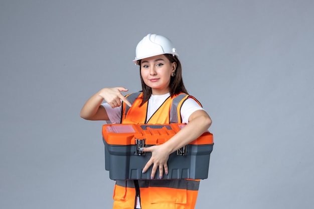 Front view of female builder holding tool case on gray wall