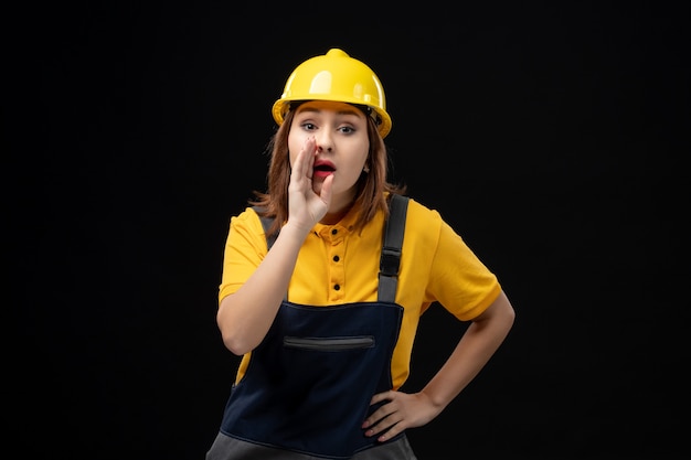 Front view female builder in uniform and helmet whispering on black wall