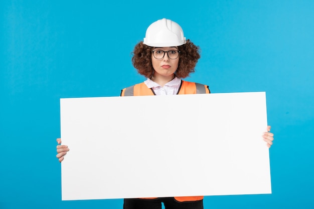 Front view of female builder in uniform holding plain desk on blue wall