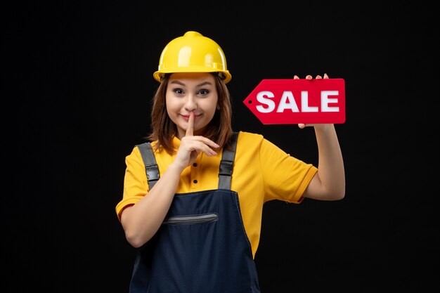 Front view female builder in uniform holding sale sign on a black wall