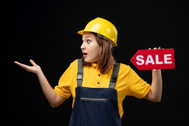 Front view female builder in uniform holding sale sign on black wall