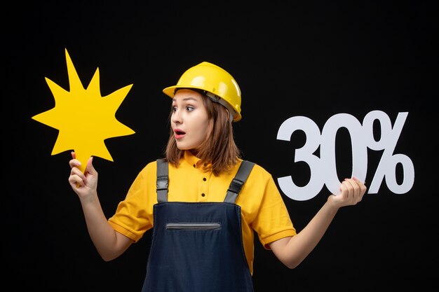 Front view female builder in uniform holding yellow figure and on black wall