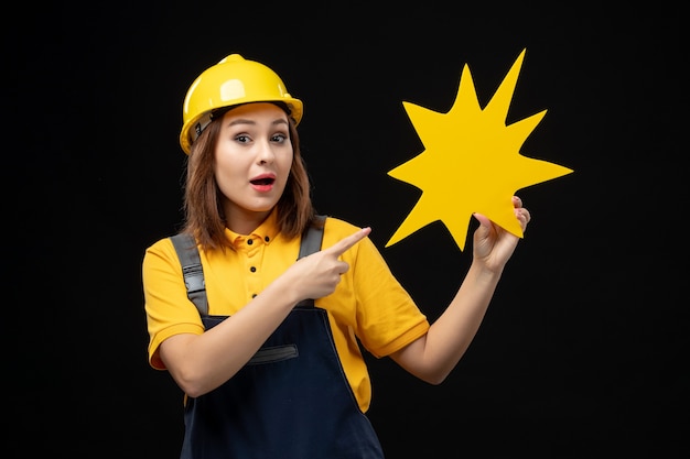 Front view female builder in uniform holding yellow figure on black wall