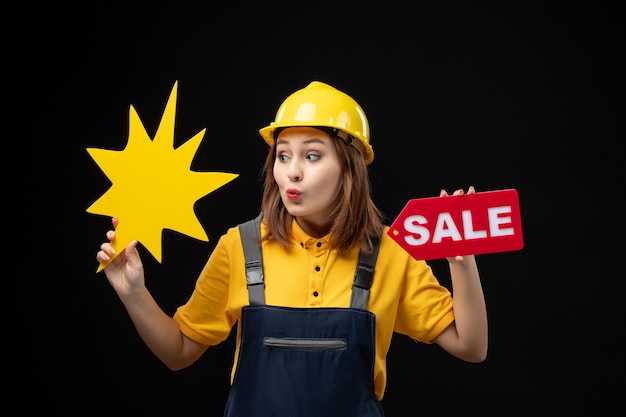 Вид спереди женщина-строитель в униформе, держащая желтую фигуру и распродажу на черной стене