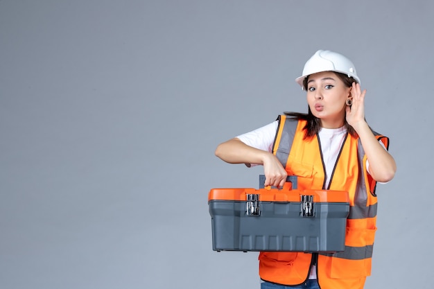 Front view of female builder with heavy tool case on gray wall