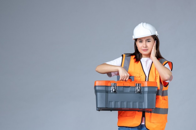 Front view of female builder with heavy tool case on gray wall