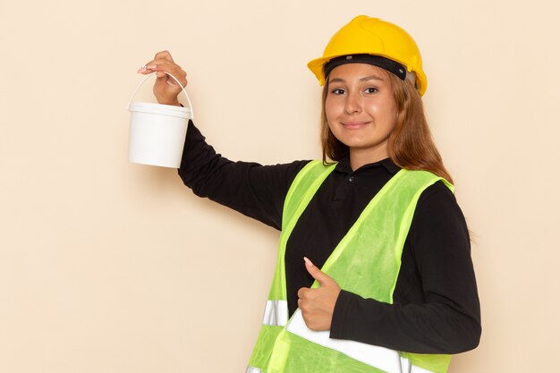 Вид спереди женщина-строитель в желтом шлеме, черная рубашка, держащая краску с улыбкой на белой стене, женщина-строитель-архитектор