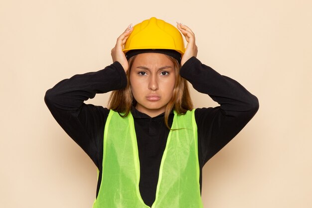 Front view female builder in yellow helmet black shirt posing having headache on the white wall 