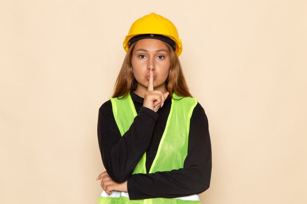 Front view female builder in yellow helmet black shirt posing showing silence sign on white wall 