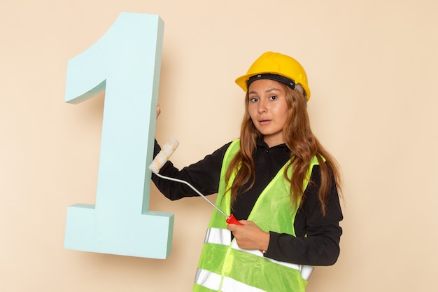 Free photo front view female builder in yellow helmet holding brush and number one figure on white wall