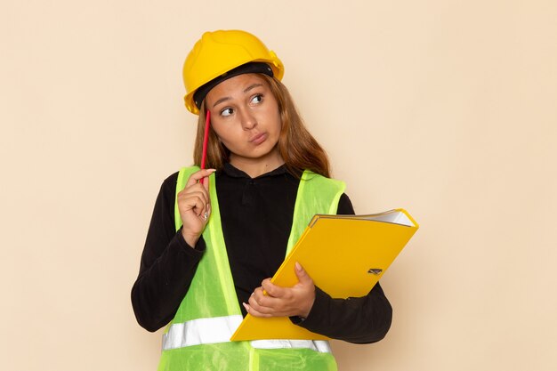 Front view female builder in yellow helmet holding yellow file and pencil thinking on white desk  