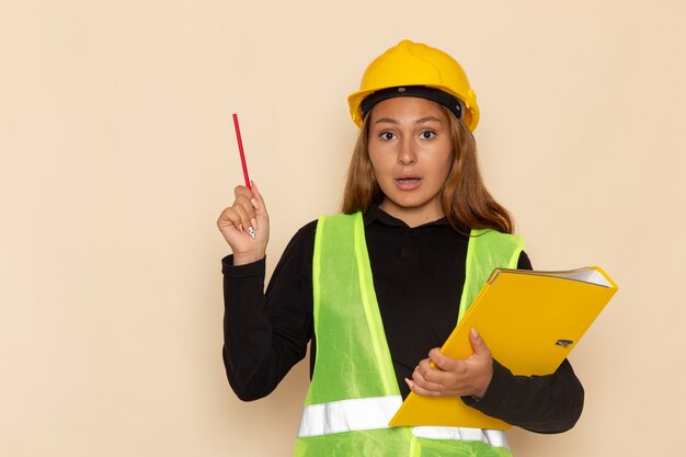 Вид спереди женщина-строитель в желтом шлеме, держащая желтый файл и карандаш на белой стене