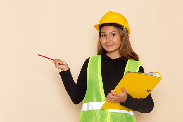 Front view female builder in yellow helmet holding yellow file and pencil on white wall  