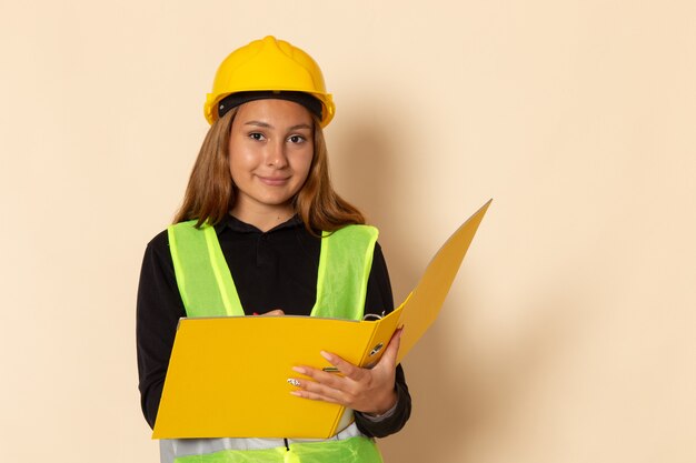 Front view female builder in yellow helmet holding yellow file writing down notes smiling on white wall  