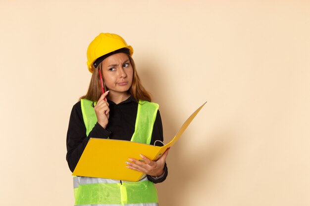 Front view female builder in yellow helmet holding yellow file writing down notes and thinking on white wall  