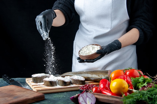 Вид спереди женщина-шеф-повар покрывает ломтики сырой рыбы мукой из свежих овощей на деревянной доске нож для муки на кухонном столе