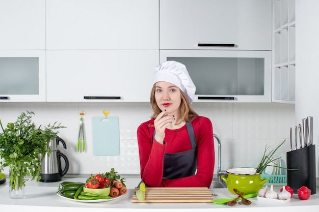 Front view female chef in uniform standing behind kitchen table putting hand on her chin