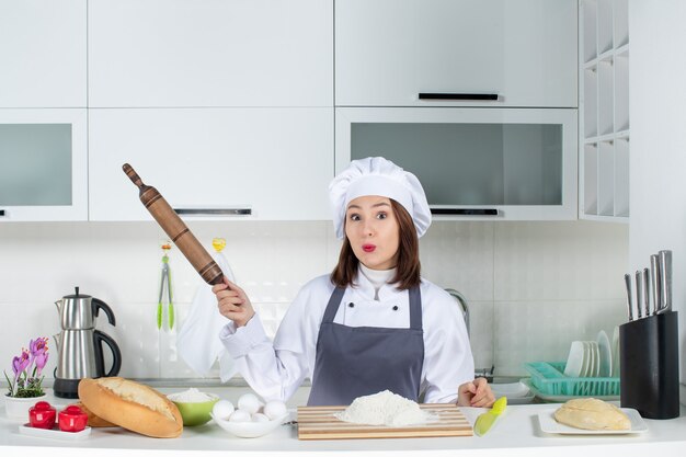 白いキッチンで麺棒を保持しているまな板の食品とテーブルの後ろに立っている制服を着た女性シェフの正面図