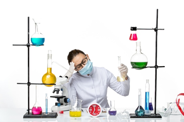 Бесплатное фото Вид спереди женщина-химик в белом медицинском костюме с маской, держащей раствор с головной болью на белом фоне, всплеск вируса covid из лаборатории химика
