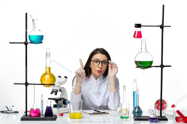 白い背景の実験室ウイルスcovidパンデミック科学の解決策とテーブルの周りに座っている医療スーツの正面図女性化学者