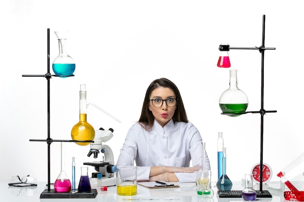 Front view female chemist in medical suit sitting around table with solutions on white background lab virus covid pandemic science