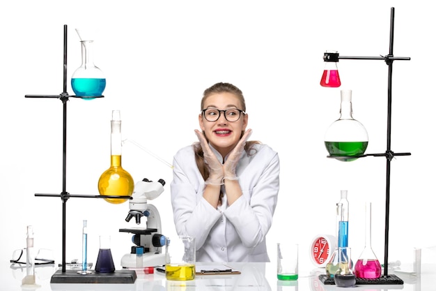 Front view female chemist in medical suit sitting with solutions and smiling on white background chemistry pandemic covid- virus