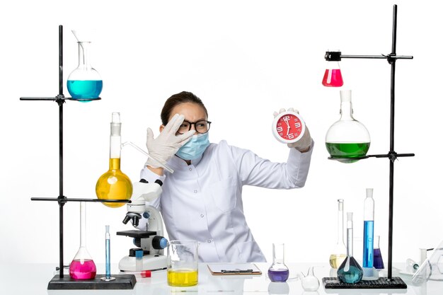 Front view female chemist in medical suit with mask holding clocks on light white background virus lab chemistry covid splash