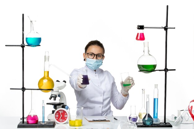 白い背景の上のさまざまなソリューションを保持しているマスクと医療スーツの正面図女性化学者スプラッシュウイルス化学実験室covid
