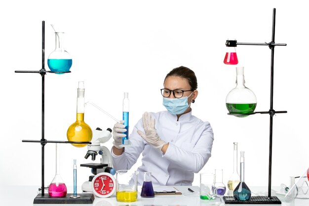Вид спереди женщина-химик в медицинском костюме с маской, держащей колбу с синим раствором на светлом белом фоне, брызги вируса, химическая лаборатория covid