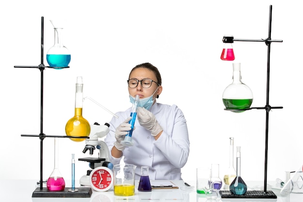 Вид спереди женщина-химик в медицинском костюме с маской, держащей колбу с синим раствором на белом фоне, брызги вируса, химическая лаборатория covid