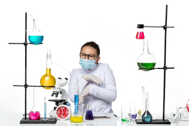 Вид спереди женщина-химик в медицинском костюме с маской, сидящая перед столом с растворами на светлом белом фоне, вирусная химическая лаборатория, всплеск covid