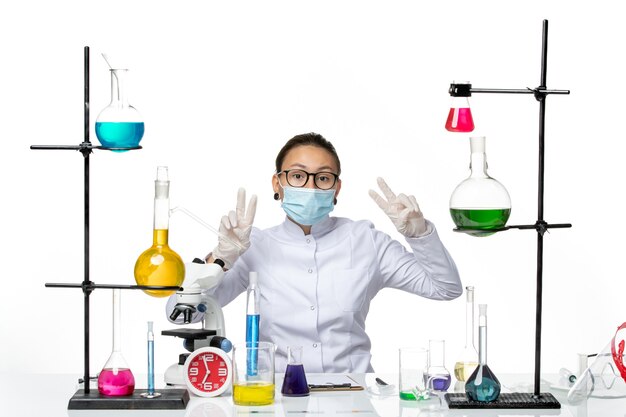 마스크는 흰색 책상 바이러스 화학 실험실 covid 스플래시에 솔루션 테이블 앞에 앉아있는 의료 소송에서 전면보기 여성 화학자