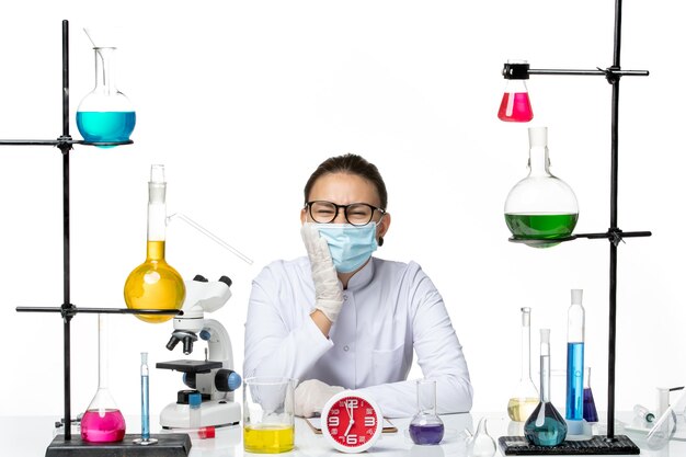 Front view female chemist in medical suit with mask sitting with solutions having toothache on white background chemistry virus lab covid- splash