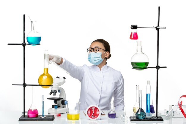 Front view female chemist in medical suit with mask sitting with solutions on light-white background chemistry lab virus covid- splash