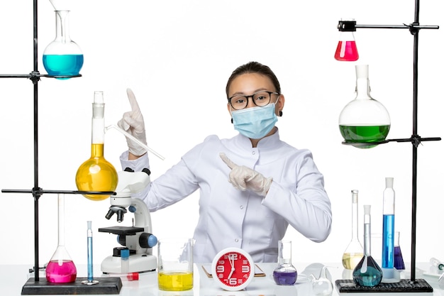 Front view female chemist in medical suit with mask sitting with solutions on light white background chemistry virus covid- splash