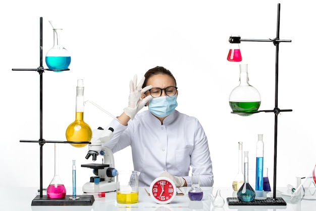 Front view female chemist in medical suit with mask sitting with solutions looking on white background chemist lab virus covid- splash