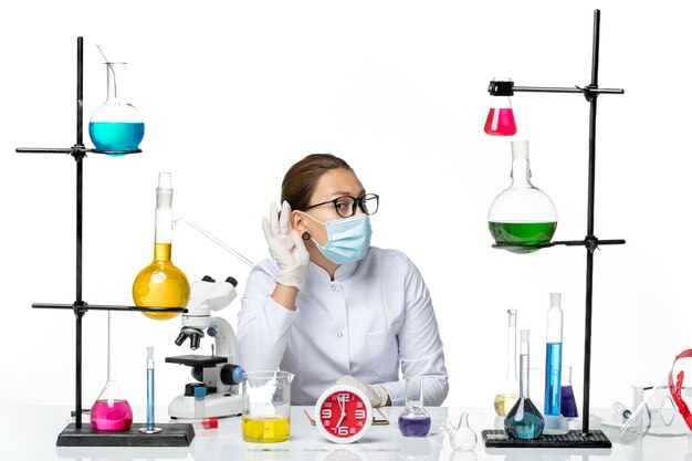 Front view female chemist in medical suit with mask sitting with solutions trying to hear on white background chemist lab virus covid- splash