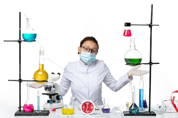 正面図白い背景の化学実験室ウイルスcovid-スプラッシュにソリューションと一緒に座っているマスクと医療スーツの女性化学者