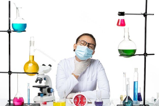 正面図白い背景の化学ウイルスラボcovid-スプラッシュにソリューションと一緒に座っているマスクと医療スーツの女性化学者