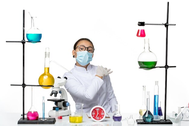 Front view female chemist in medical suit with mask sitting with solutions on a white background splash virus chemistry covid- lab