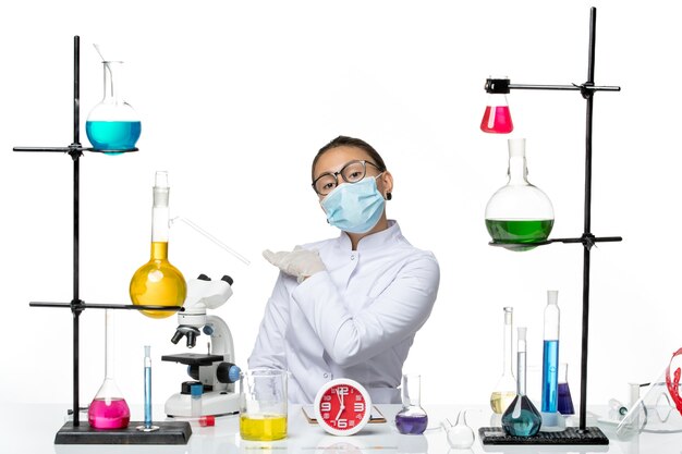 Вид спереди женщина-химик в медицинском костюме с маской, сидящая с растворами на белом фоне, всплеск вируса, химия, лаборатория covid