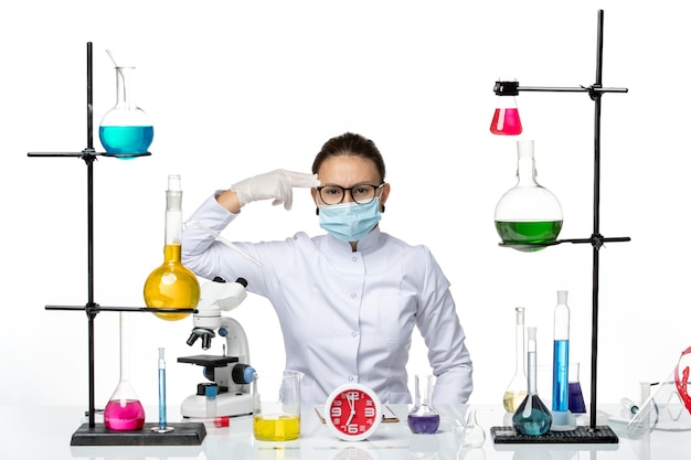 Вид спереди женщина-химик в медицинском костюме с маской сидит с растворами на белом столе химическая вирусная лаборатория covid- splash