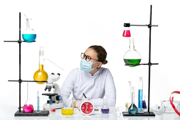 正面図白い背景の化学者ラボウイルスcovid-スプラッシュにメモを書くソリューションと一緒に座っているマスクと医療スーツの女性化学者